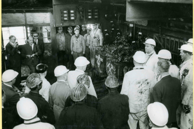 Ansprache eines Grubendirektors vor Bergleuten und Journalisten mit geschmückten Grubenwagen. 