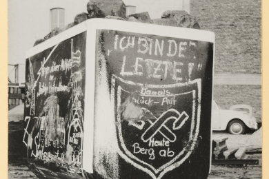 Kreidezeichnungen auf einem Grubenwagen mit verbogenem Schlägel und Eisen-Symbol und Aufschrift „Ich bin der Letzte! Damals Glück-Auf, Heute Berg ab“.