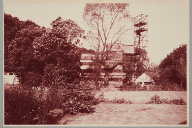 Hinter Gärten im Wald stehendes stählernes Gerüst und ein Gebäude sowie ein brückenartiges Gebilde