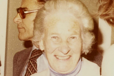 Ausschnitt aus einer Fotografie, der eine lachende, ältere Frau in hellem Kostüm und mit einem Sektglas in der Hand zeigt.