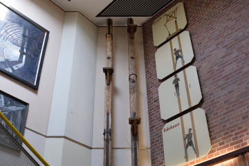 Zwei hölzerne, sechs Meter lange Balken mit Tritten und Griffen an der Wand eines Treppenhauses im Deutschen Bergbau-Museum Bochum, daneben Tafeln mit Bildern, die zeigen, wie ein Bergmann sich auf den Balken hin und her bewegt, um im Schacht nach oben oder unten zu fahren.