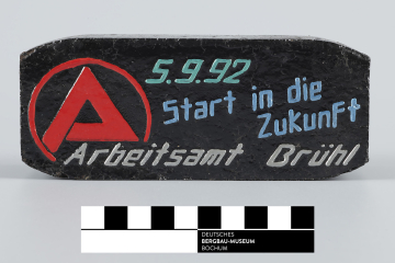 Das Foto zeigt ein mehrfarbiges Schmuckbrikett. In rot ist das Symbol des Arbeitsamtes gehalten, grünlich das Datum, der 05.09.1992, bläulich der Satz „Start in die Zukunft“ und silbern Arbeitsamt Brühl.