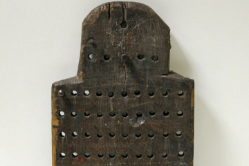 Rechteckiges Holzbrett im Hochformat, oben halbrund, versehen mit je zehn Löchern in zehn Reihen und zwei Holzpins zum Einstecken. Im halbrunden Bereich befinden sich weitere fünf Löcher in Reihe.