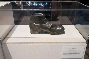 Schwarzer Lederstiefel, ausgestellt unter einer Acrylglashaube, auf dem Sockel ein Schild mit der Objektbeschreibung