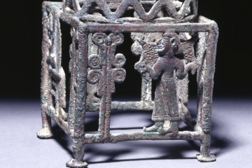 Vierseitiger, stark korrodierte Ständer, auf einer der Seiten Kupferbarrenträger mit geschultertem Ochsenhautbarren, in Richtung eines stilisierten Baumes sehend