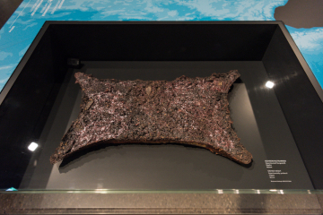In einer Vitrine liegender, in der Form an eine gespannte Rinderhaut erinnernder Kupferbarren mit unebener Oberfläche