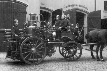 Das Foto zeigt ein von Pferden gezogenes Löschfahrzeug mit Feuerwehrleuten 