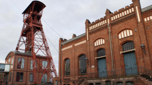 Ein Fördergerüst steht neben einem großen Backsteingebäude mit Bogenfenstern.